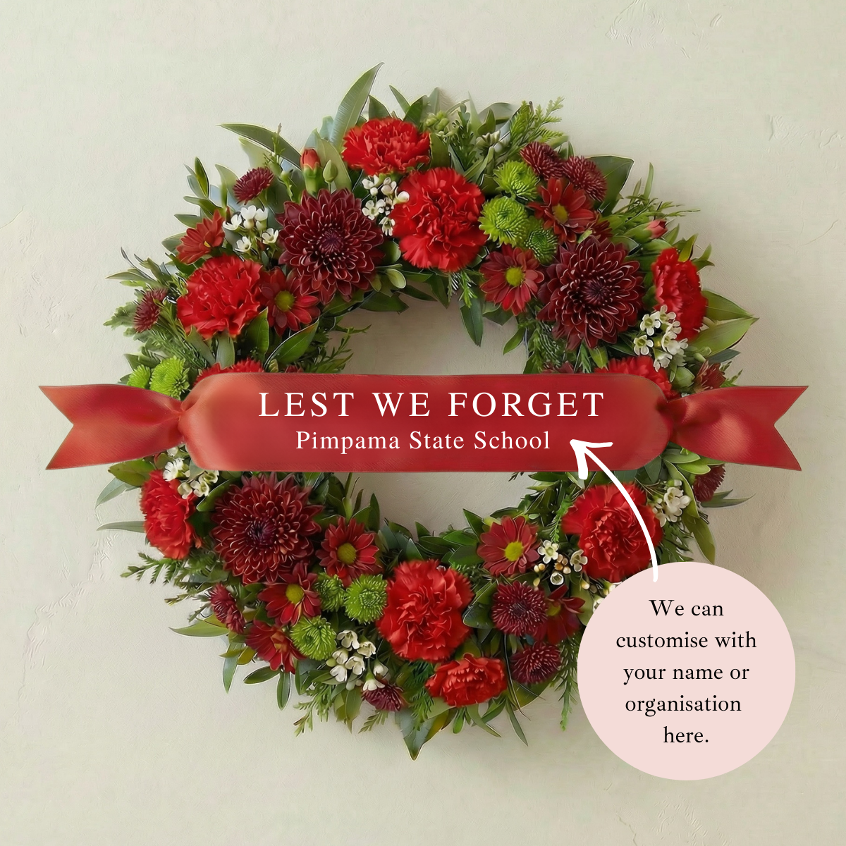 ANZAC remembrance wreath with red flowers and greenery featuring a custom satin ribbon reading “Lest We Forget – Pimpama State School”.